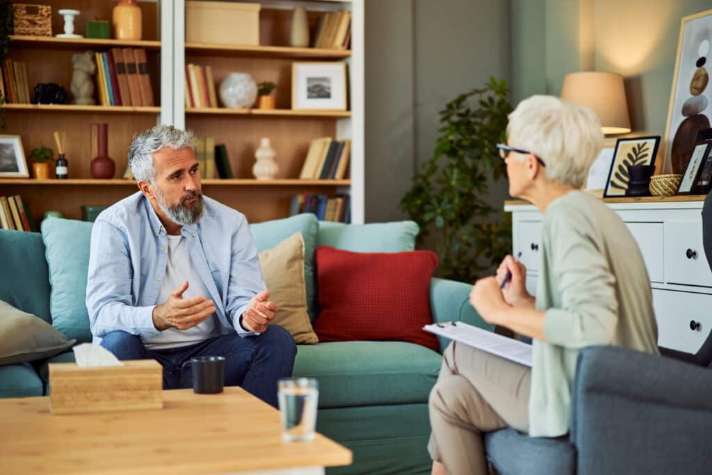 A mentally exhausted senior adult man explaining his problems to his psychotherapist while sitting on a couch in the doctor's office.