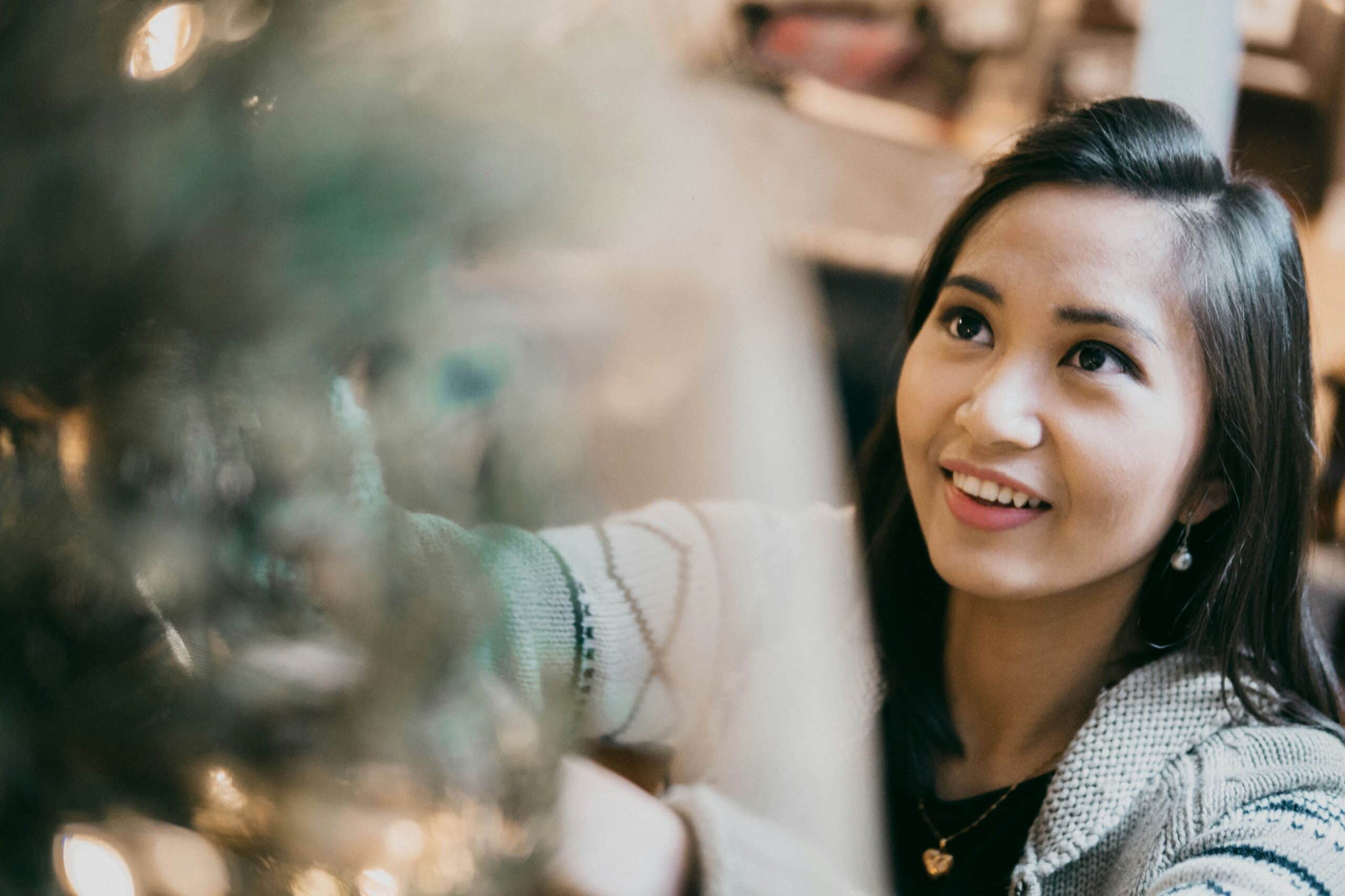 A woman is smiling while decorating a Christmas tree, gazing up at the ornaments. She is wearing a cozy sweater and delicate jewelry, and her expression is joyful and focused. The photo is slightly blurred with twinkling lights in the background, creating a warm, festive atmosphere.