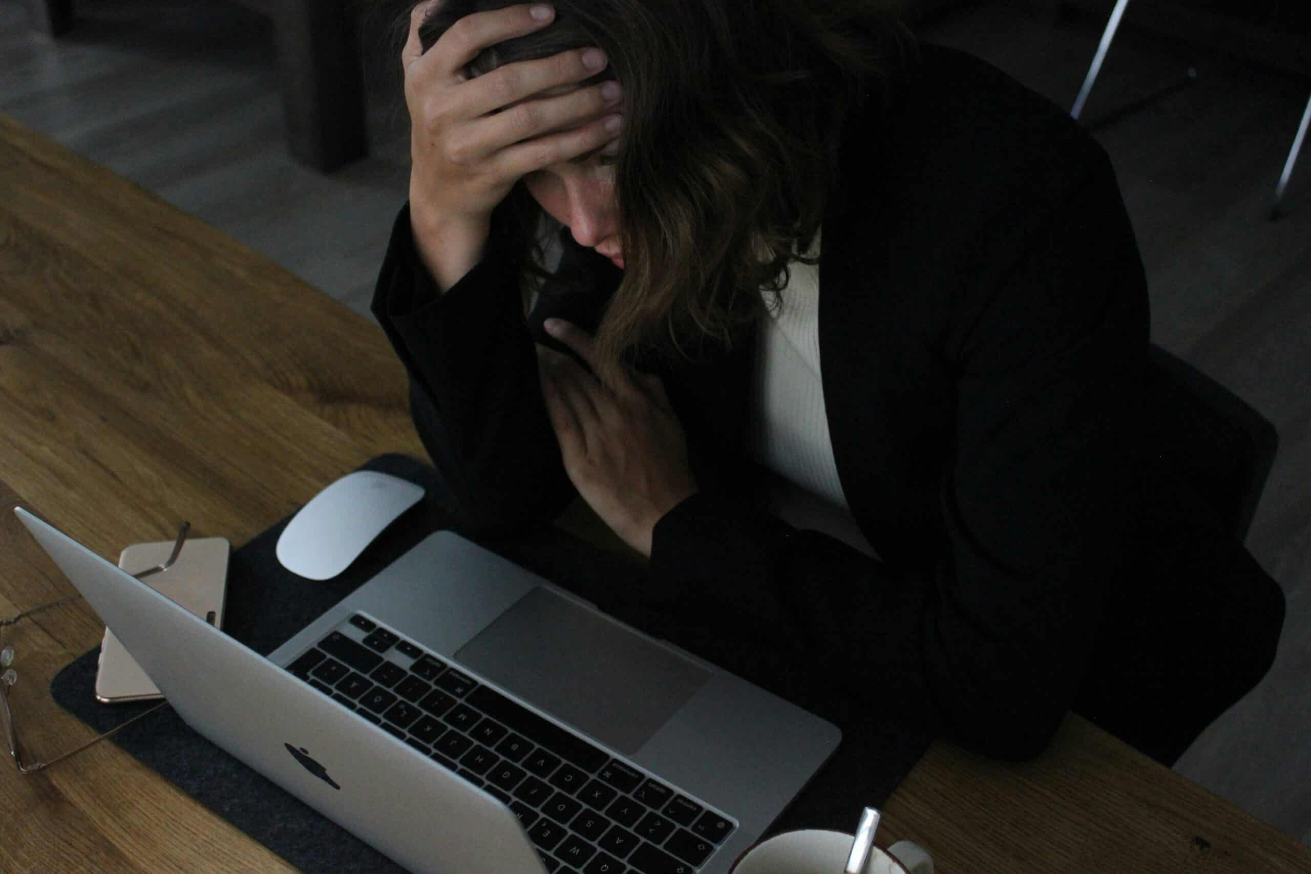 woman in black jacket bent over laptop with hand on her face